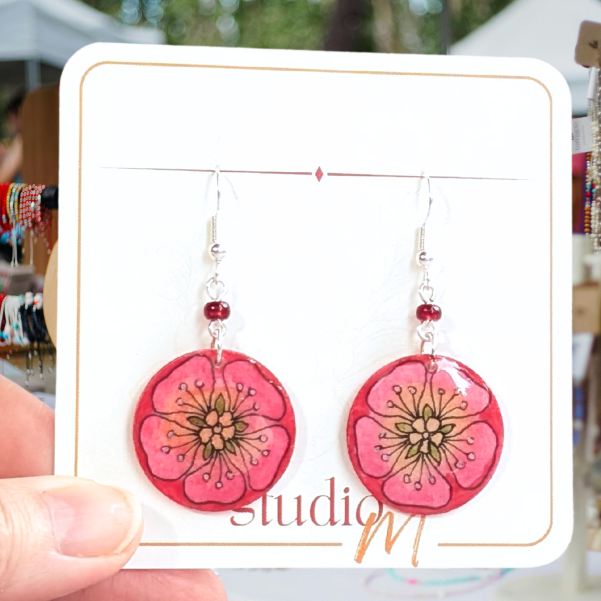A pair of dangling earrings with round pink flower charms and silver hooks, displayed on a white card held by a hand in an outdoor market setting.