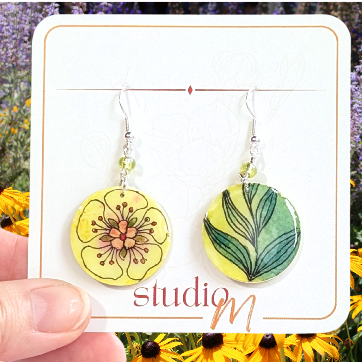 A pair of round dangle earrings displayed on a white card, held by a hand against a background of yellow flowers and purple lavender. The left earring features a yellow flower with black and orange details, and the right earring features green leaves on a yellow background.