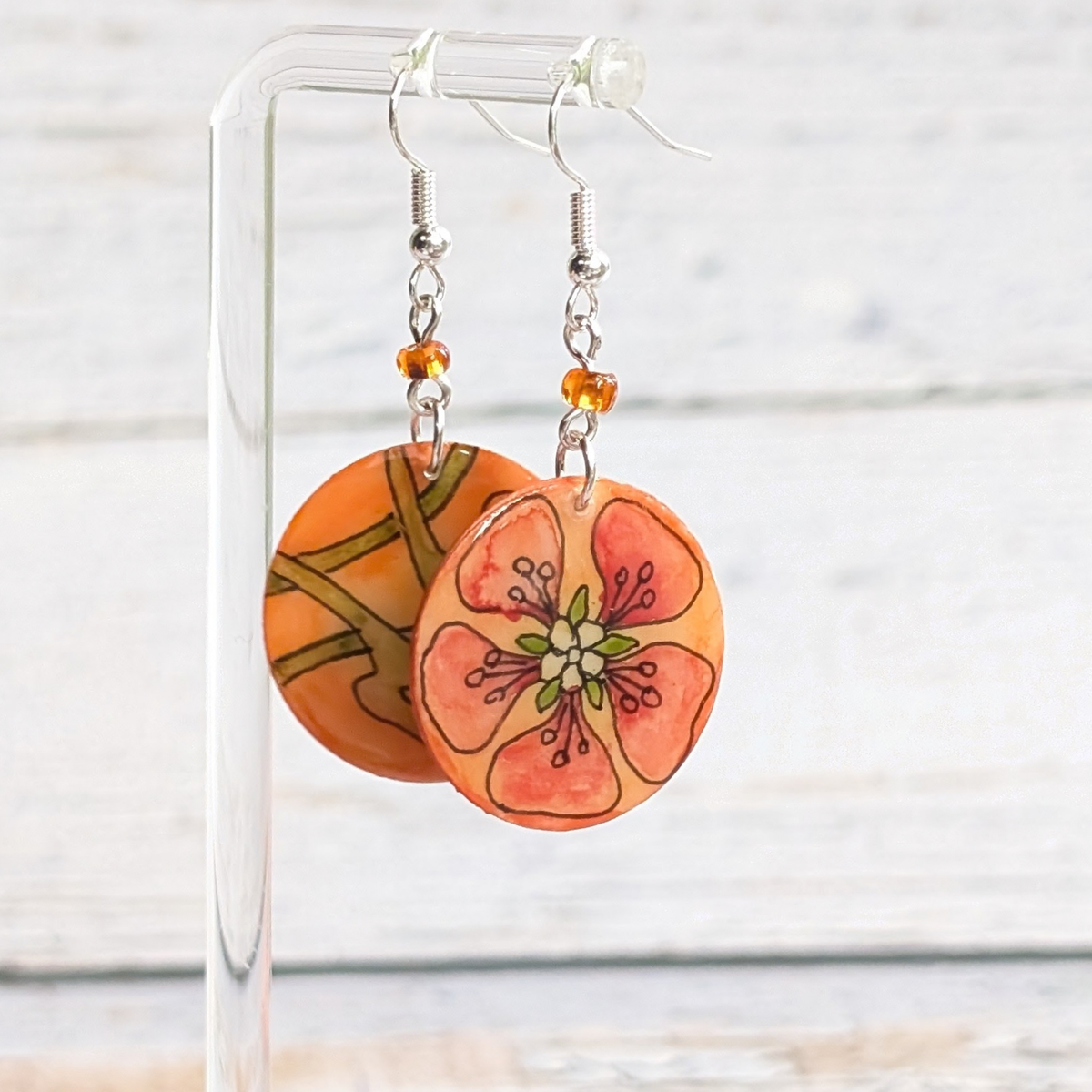A pair of handmade dangle earrings with circular orange and peach flower charms, hanging from silver fishhook wires with small amber beads, displayed on a clear acrylic stand against a white wood background.