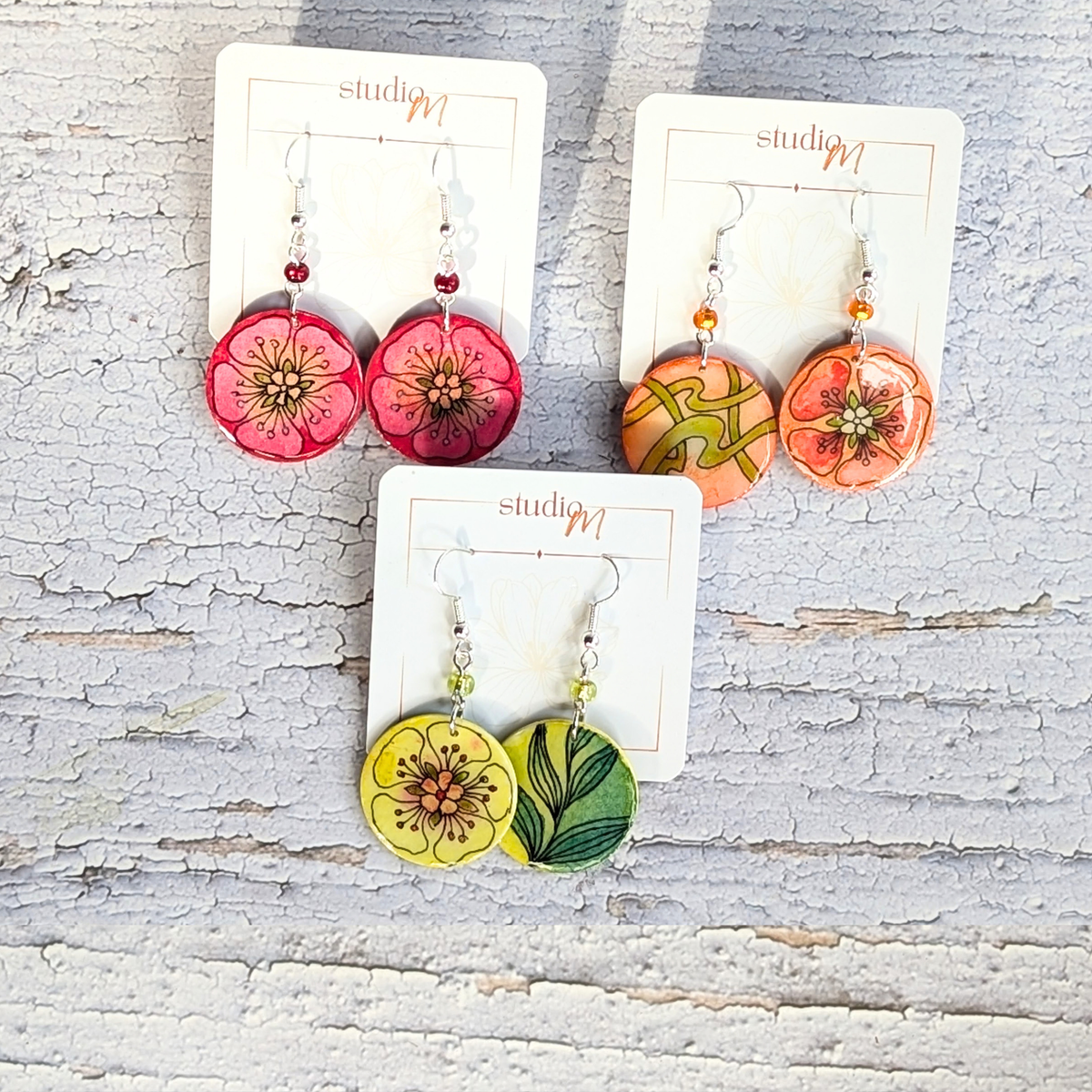Three pairs of floral dangle earrings displayed on white card holders against a distressed white wood background. The earrings feature round flower-shaped charms in pink, orange, and yellow with green and brown floral details, connected by small beads.