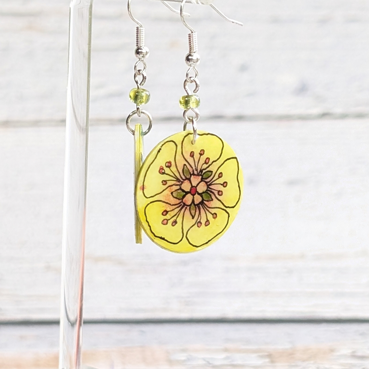 Yellow flower-shaped dangle earrings with silver hooks and small green beads, displayed on a clear stand against a light wood background.