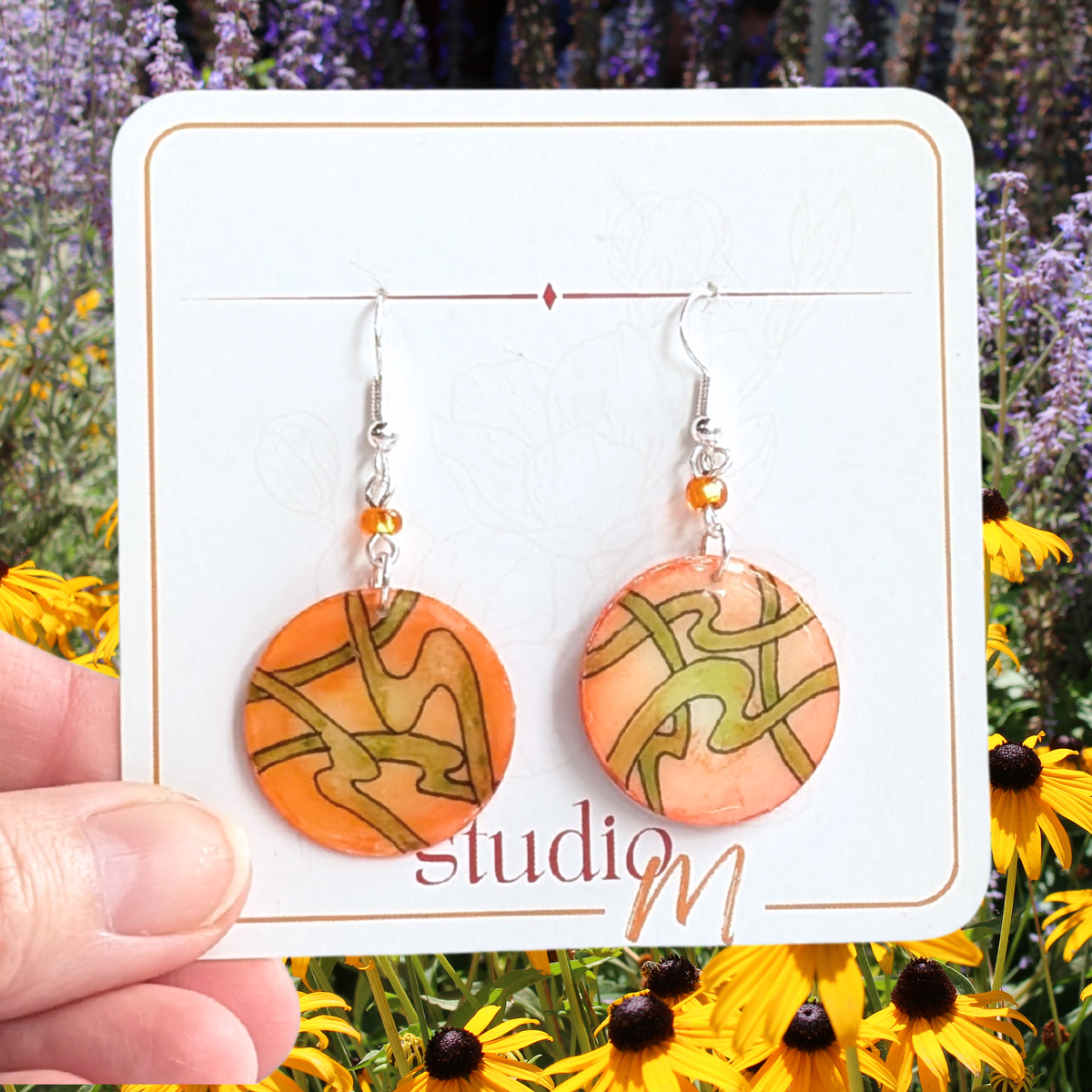 A pair of round orange and peach drop earrings with olive green abstract line patterns, hanging from silver hooks with small orange beads. They are displayed on a white card held by a hand against a background of yellow flowers and purple lavender.
