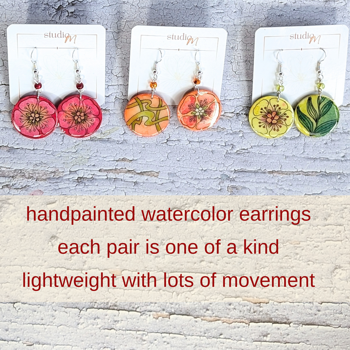 Three pairs of handpainted watercolor dangle earrings displayed on white cards against a distressed white wood background, with red text below reading 'handpainted watercolor earrings each pair is one of a kind lightweight with lots of movement.' The earrings feature floral and botanical designs in pink, orange, and green tones.