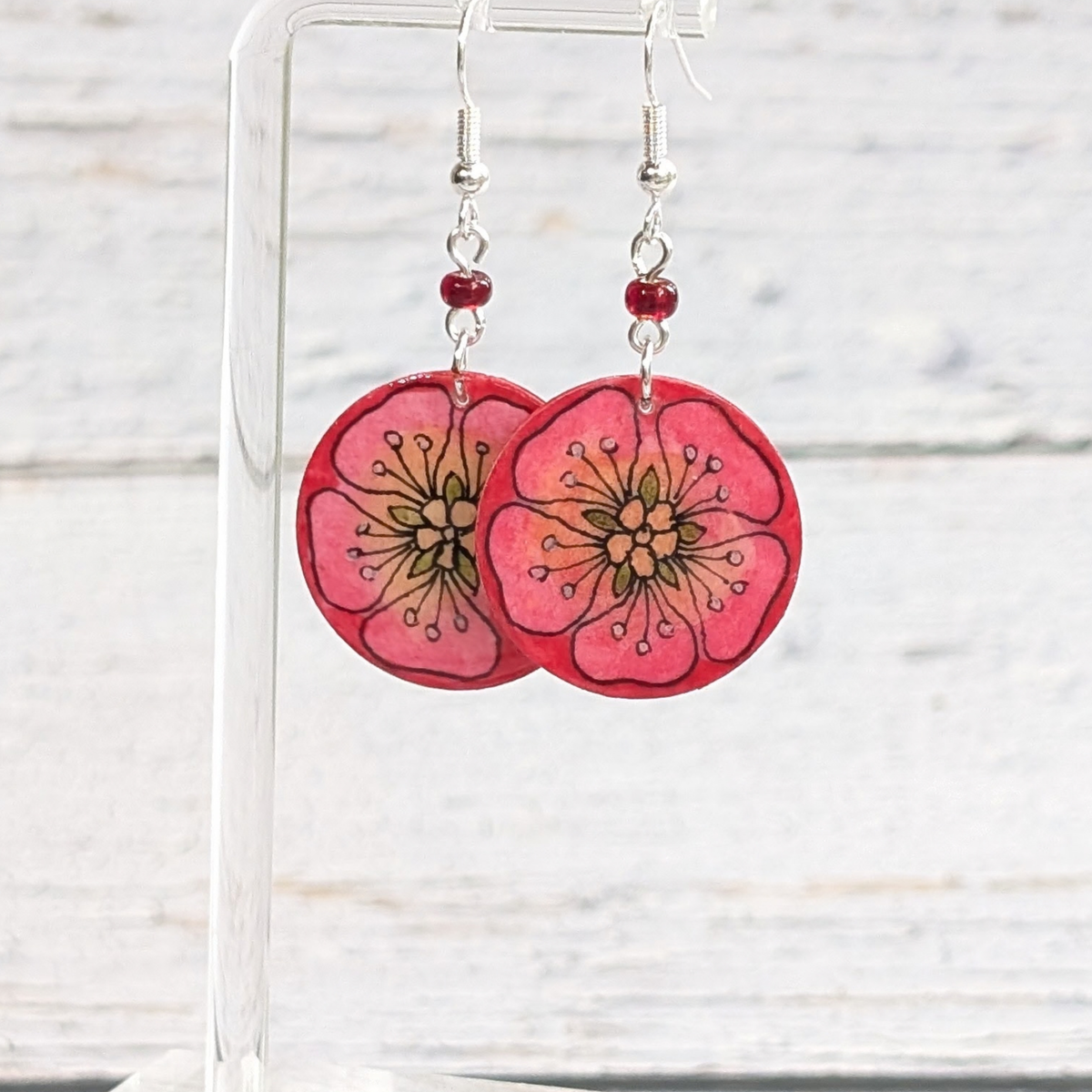 A pair of handmade dangle earrings featuring pink cherry blossom flower charms with black line details, hanging from silver hooks with small red beads, displayed on a clear acrylic stand against a white wood background.